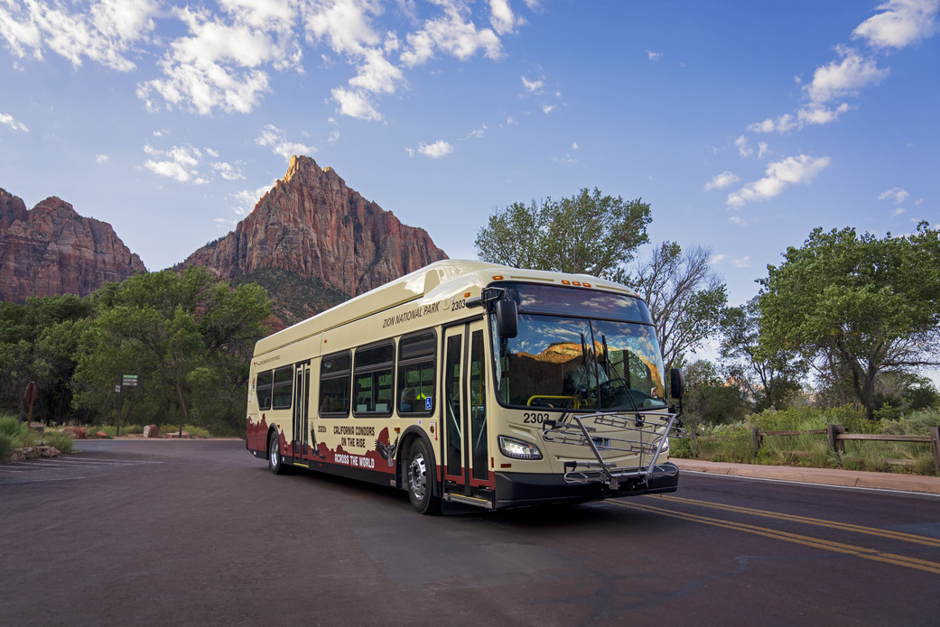 Picture of a full-size Zion National Park Bus on the road with the mountains of the park in the background.  On the side it says "California condors on the rise across the world".